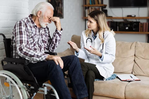 A healthcare worker talks to a patient. The patient, an older man, is sitting in a wheelchair.