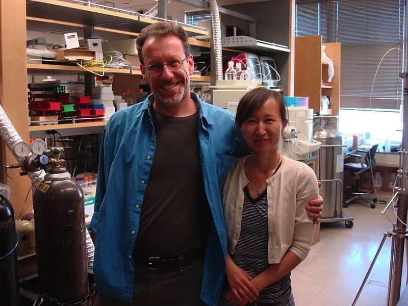 Dr. Bitan stands with an arm around a researcher. They are smiling. Metal tanks, pressure gauges, and tubes are visible in the background.