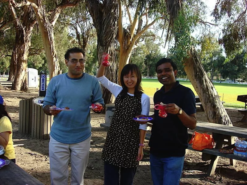 Three lab members hold paper plates with food and candy canes during a day at the park. Trees and wooden picnic tables are visible behind them..