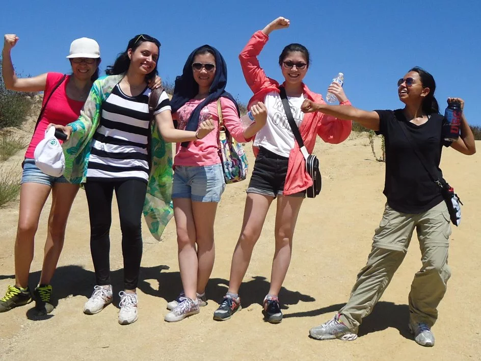 Six students stand on a sandy dune, flexing their arms on a sunny day.