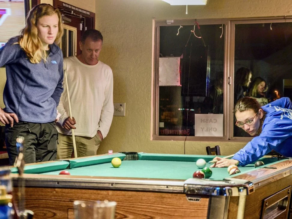 Two lab members watch another student finish his turn at pool. 