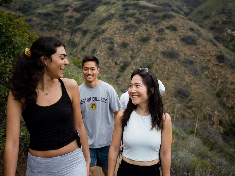Two of the lab's female students are facing each and smiling during a hike. A male student is walking behind them, also smiling. 