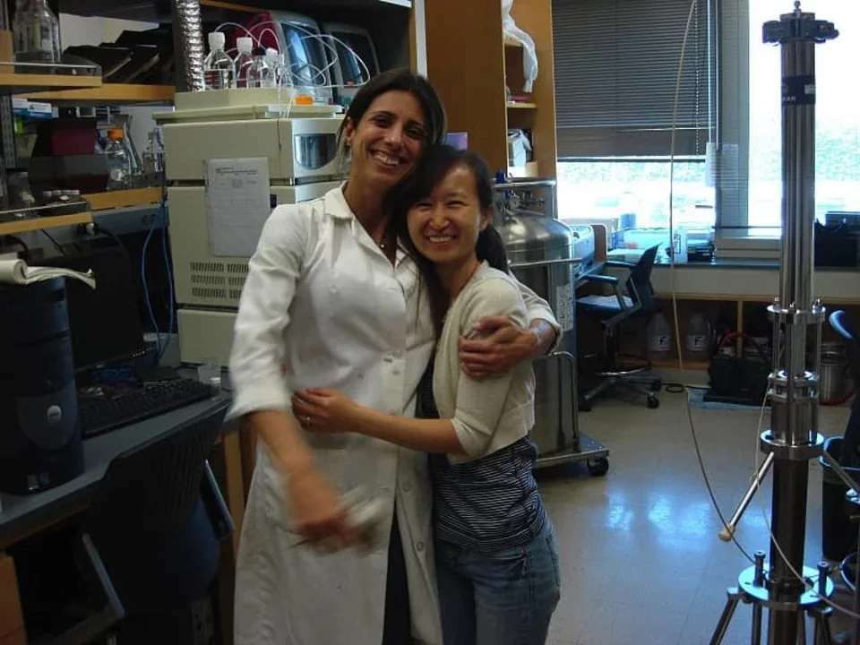 Two researchers, one in a white lab coat, are standing in the lab with their arms around each other, laughing.