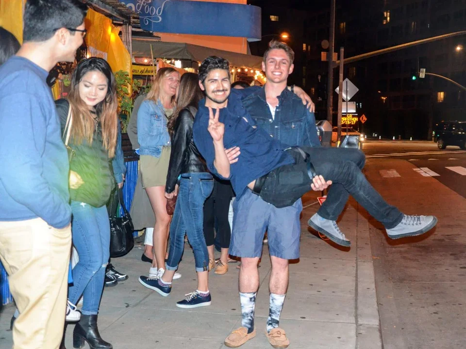 A lab student carries another student outside of a restaurant. Other students are having conversations in the background. 