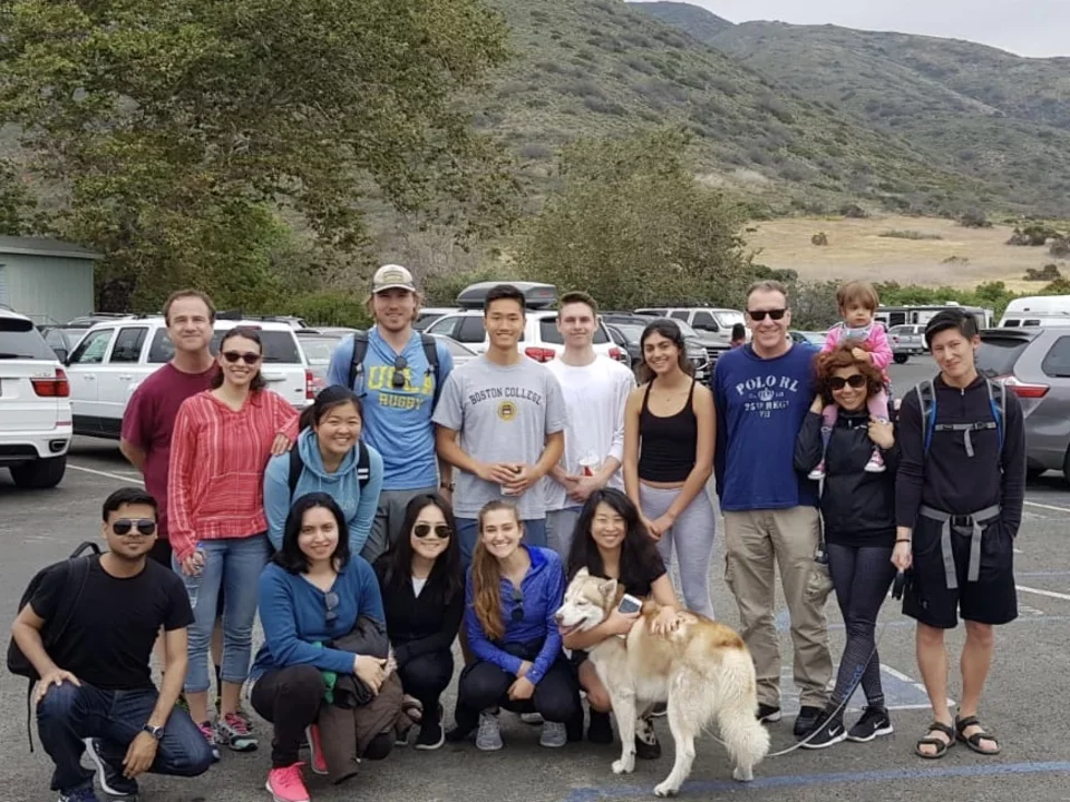 A large group of students stand in a parking lot. A second row of students kneel in front of them. Everyone is looking at the camera and smiling.