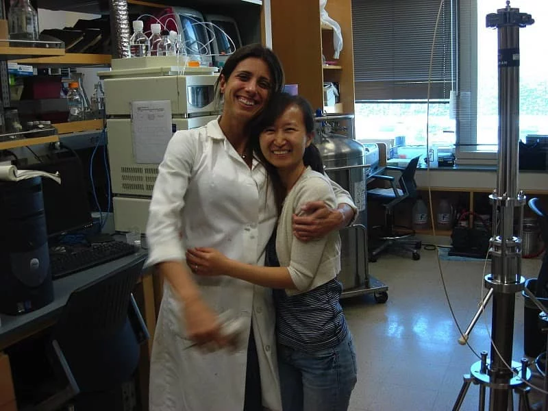 Two researchers, one in a white lab coat, are standing in the lab with their arms around each other, laughing.